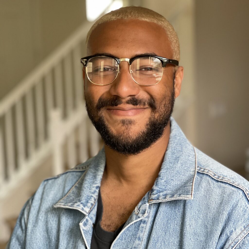 A photo of Justin Warren in classic glasses, blonde hair, and a faded denim jacket. 